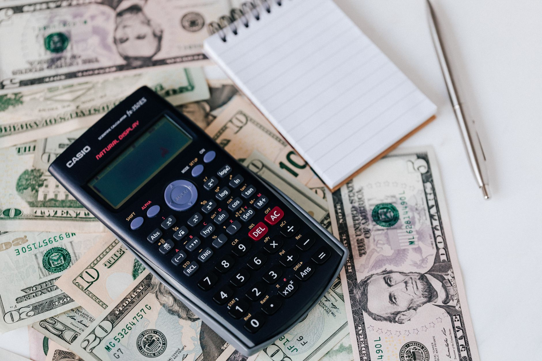 A calculator, US dollar bills, and a notepad on a desk — tallying the cost of duplicate payments